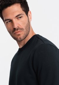 Sudadera negra con cuello redondo, con puños y dobladillo de canalé, textura suave y un sutil detalle de costura en el hombro.