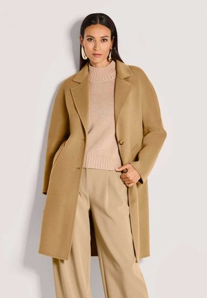 Vrouw in een oversized camel kleurige jas over een beige gebreide trui en wijde broek, accessoires met grote gouden oorbellen en een ring.