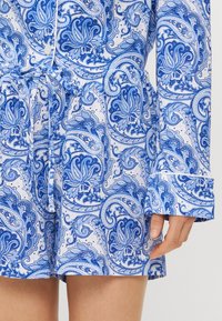 Blue and white patterned romper featuring paisley design, drawstring waist, long sleeves, and buttoned cuffs. Soft fabric texture.