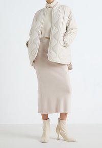 Veste rembourrée beige avec motif en vagues, pull en maille crème à col roulé, jupe ajustée beige clair et bottines crème avec détail de fermeture éclair.