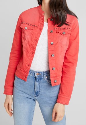 Vrouw draagt een fel koraalkleurige denimjas met knopen en metalen studs op de borstzakken, een wit shirt en lichtblauwe spijkerbroek.