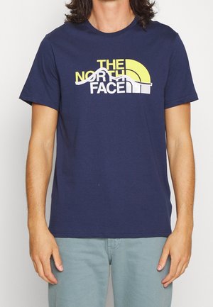 Henkilö pukeutunut tummansiniseen t-paitaan, jossa on keltavalkoinen "The North Face"-logo ja vaaleansiniset housut.