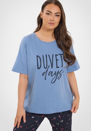 Mujer con cabello largo y castaño vistiendo una camiseta azul claro con el texto "DUVET days" y pantalones de pijama oscuros con pequeños patrones coloridos.