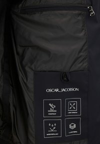 Sort jakke med en blank overflade, der har en lynlåslomme, brandlogo og ikoner, der angiver vindafvisende, vandafvisende og strækegenskaber.