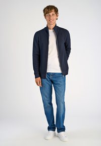 Marineblauer, gestrickter Zip-Jacke mit Rippstruktur, darunter ein weißes T-Shirt, blaue Jeans und weiße Sneaker. Stehende Figur mit den Händen in den Taschen.