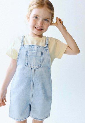 Fille rousse souriante avec des taches de rousseur, portant un t-shirt jaune et une salopette en denim bleu clair, touchant son oreille d'une main.