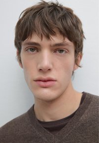 Portrait of a young male with short, tousled brown hair and green eyes. He wears a brown V-neck sweater over a dark shirt. Smooth skin texture.