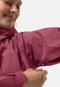Giacca impermeabile rosa con finitura testurizzata, dotata di tasca con zip e polsini regolabili. Marchio visibile sul lato sinistro.