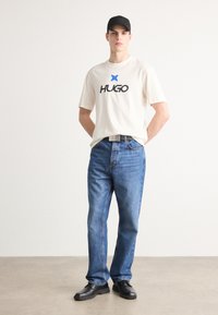 Beige cotton t-shirt with black "HUGO" logo, blue star; blue denim jeans; black cap; black shoes. Model stands with hands on hips.