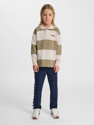 Kind met lang blond haar, gekleed in een beige-wit gestreept poloshirt, marineblauwe trainingsbroek en witte sneakers, staand tegen een effen achtergrond.