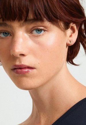 Gold crescent-shaped stud earring with textured detail, worn on the right ear. Close-up of facial features with neutral background.