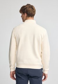Suéter blanco de algodón con cuello alto, puños acanalados y dobladillo. Textura suave y diseño sencillo, combinado con pantalones oscuros.