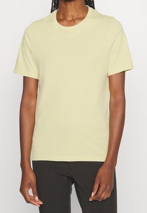 Person wearing a pale yellow short-sleeve crew neck t-shirt paired with black pants, standing against a plain light background.