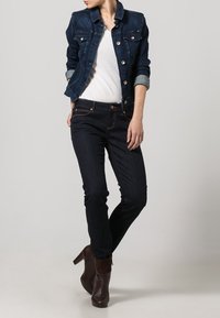 Giacca di denim scuro con bottoni metallici, t-shirt bianca sotto, abbinata a jeans scuri e stivaletti marroni. Design semplice e aderente.