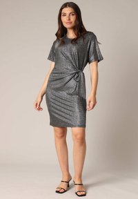 Robe grise scintillante à manches courtes, détail noué à la taille et silhouette ajustée, accompagnée de sandales noires à lanières.