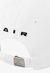 Casquette blanche avec lettrage "AIR" brodé en noir. Présente une texture douce en coton et une fermeture à boucle en métal argenté avec un accent de logo.