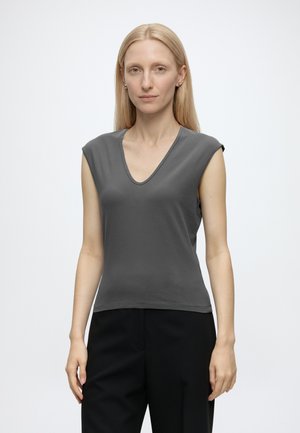 Femme aux cheveux blonds et lisses, debout face à l'objectif, portant un haut gris sans manches à col en V et un pantalon noir sur un fond uni clair.