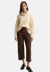 Kobieta stoi w kremowym swetrze z golfem, brązowych, krótkich spodniach w szerokim kroju oraz czarnych butach sznurowanych na jednolitym tle.