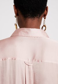 Ljust rosa satinblus med krage, slät textur, bakfåll och en söm vid nederkant. Presenterade är örhängen i guldton.