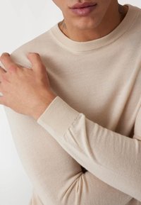 Beiger Strickpullover mit Rundhalsausschnitt und gerippten Bündchen. Nahaufnahme der Stoffstruktur und Hände, die über die Schulter kreuzen.