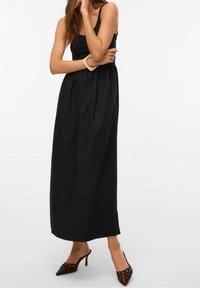 Mujer vistiendo un vestido maxi negro sin mangas, pulsera beige y mules de tacón marrón con puntera afilada y ojales decorativos.