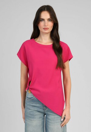 Femme aux longs cheveux foncés portant un haut rose vif à manches courtes avec un ourlet asymétrique et un jean bleu sur fond gris.