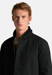 Sort jakke med en ståkrave, der har knapper og en glat, elegant tekstur. Bæres over en lysere sweater.