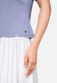 Hellbluse mit kurzen Ärmeln und ausgestellt geschnittenem Saum, der am unteren Ende mit metallicfarbenem Besatz verziert ist. Weißer Rock mit vertikalen silbernen Streifen.