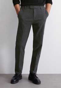 Pantaloni grigi su misura con design strutturato, davanti piatto, tasche laterali e tessuto leggermente strutturato. Abbinati a scarpe nere.