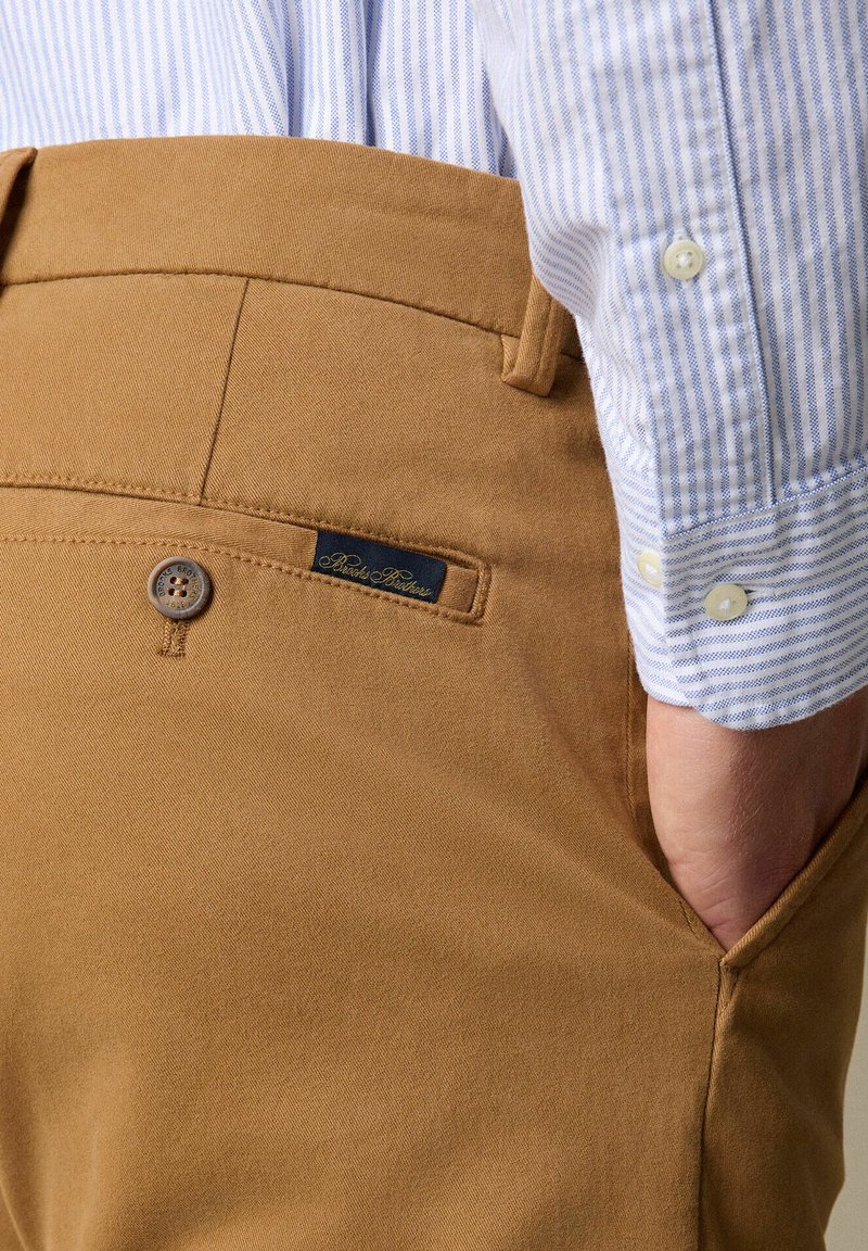Beige Baumwollhose mit glatter Textur, ausgestattet mit einer Vordertasche, Knopfverschluss und einem schwarzen Etikett-Detail. Hellbluse gestreifte Hemd sichtbar.