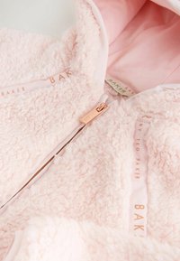 Jasnoróżowa, puszysta kurtka z polaru z miękkim podszyciem, z różowozłotym zamkiem błyskawicznym i markową taśmą wzdłuż krawędzi.