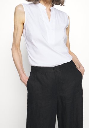 Person trägt eine ärmellose weiße Bluse, die in hoch taillierte, locker sitzende schwarze Hosen gesteckt ist, mit den Händen in den Taschen, vor einem einfarbigen Hintergrund.