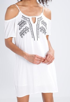 Kvinde iført en hvid cold-shoulder kjole med sort geometrisk broderi på brystet, stående med hænderne blidt foldet.