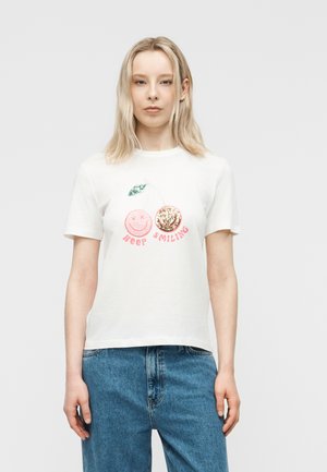 Mujer rubia con camiseta blanca que lleva dos cerezas, una rosa con cara sonriente y otra con lentejuelas doradas, y las palabras "keep smiling" en rosa.