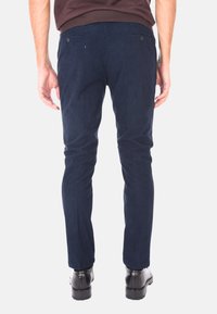 Uomo che indossa pantaloni a righe blu navy e scarpe eleganti nere, in piedi con le mani rilassate ai lati, vista da dietro.