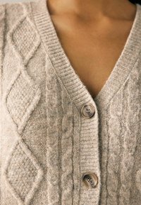 Heledbeez värvi vahvikeelne kardigan, millel on sügav V-kaelus ja kaks ümmargust nupuvajutust. Tekstuurilised mustrid ja ribakujulised varrukad lisavad detaili.