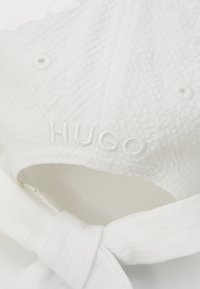Gorra blanca con tejido texturizado, logo bordado "HUGO" en hilo blanco y una correa de tela atada en la parte trasera.