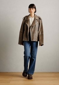 Bruine leren motorjack met brede revers, ritssluiting en oversized fit. Gecombineerd met blauwe jeans en donker gemusterde schoenen.