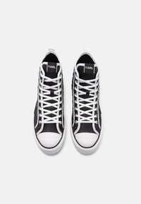 Vysoké tenisky s černým textilním svrškem, bílými tkaničkami a gumovou špičkou. Na jazyku jsou viditelné texturované detaily a logo.