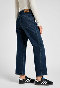 Tummansiniset farkut, suora lahje, takana ruskea logo-patch. Yhdistetty mustiin ja valkoisiin lenkkareihin.