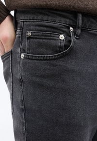 Κοντινό πλάνο χεριού ατόμου στην τσέπη μαύρου τζιν με εμφανείς ραφές και ασημένιες ειδικές διακοσμητικές βίδες.