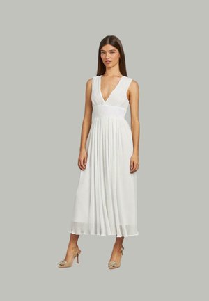 Vestido blanco sin mangas con un escote en V profundo, falda plisada y cintura ajustada. Combinado con zapatos de tacón alto beige.