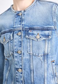 Chaqueta de denim azul claro con cierre de botones en el frente, dos bolsillos en el pecho y un escote redondeado. Presenta un desgaste sutil y detalles en costuras color canela.