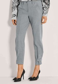 Grijze op maat gemaakte broek tot de enkel met voorplooien en kleine gespdetails aan de manchetten, gecombineerd met zwarte hoge hakken met grote strikken.
