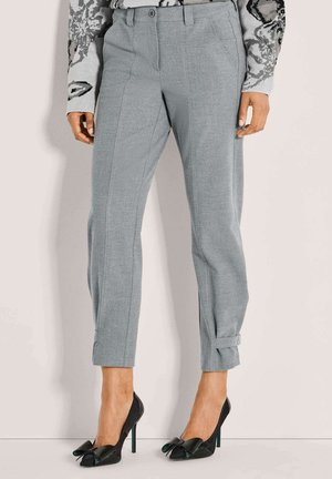 Grijze op maat gemaakte broek tot de enkel met voorplooien en kleine gespdetails aan de manchetten, gecombineerd met zwarte hoge hakken met grote strikken.