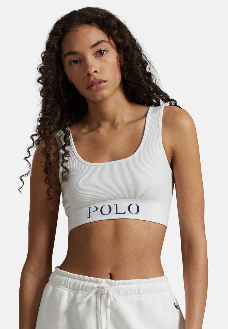 Bílý žebrovaný crop top s širokými ramínky, s námořnicky modrým logem "POLO" na elastickém pásu. Hladká textura, přiléhavý střih.