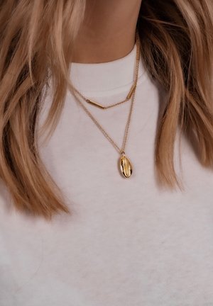 Zlaté vrstvené náhrdelníky s pendantom v tvare mušle cowrie a geometrickými prízvukmi, umiestnené na svetlej texturovanej tkanine.