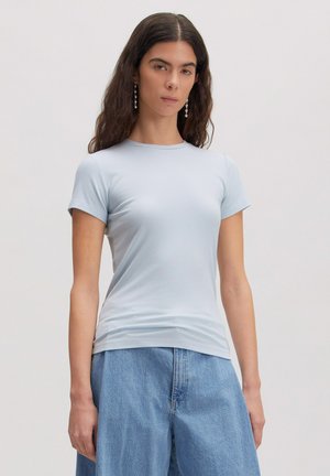 Frau mit langen dunklen Haaren, die ein hellblaues eng anliegendes kurzärmeliges Oberteil und lockere blaue Jeans trägt, steht vor einem einfarbigen Hintergrund.