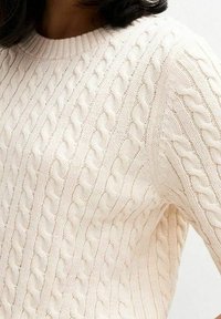 Nainen, joka on pukeutunut vaaleanvaaleaan palmikkoneuleeseen, jossa on pyöreä kaula-aukko. Neuleessa näkyy yksityiskohtainen rakenne ja neuloskuvio.