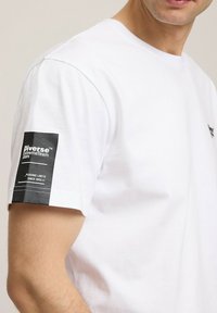 Biała bawełniana koszulka z krótkim rękawem, z czarną tkaninową łatą na lewym rękawie z tekstowym brandingiem i białym detalem w stęgu.